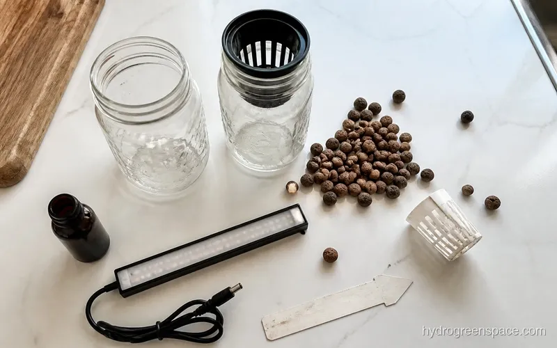 Overhead flat lay of Kratky starter kit with mason jars, net cups, clay pebbles, dark nutrient bottle, and LED bar on a white counter