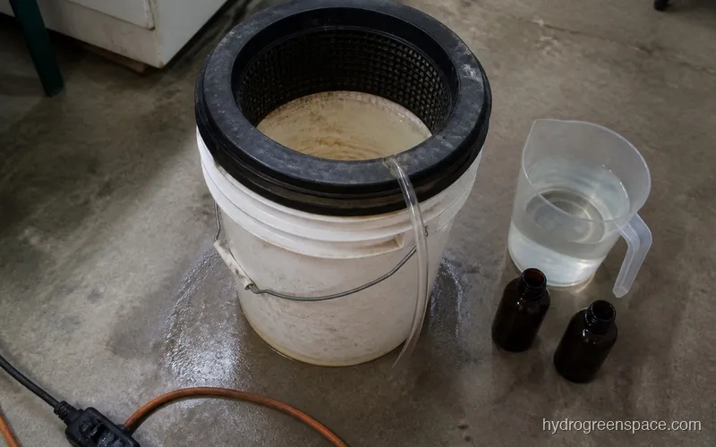 Hydroponic DWC bucket with reservoir being drained and refilled next to a measuring cup and nutrient bottles